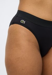 Μαύρο κάτω μέρος μπικίνι με φαρδύ ζωνάρι, με μικρό κεντητό λογότυπο κροκοδείλου στο πλάι. Το υλικό φαίνεται λείο και ελαστικό.