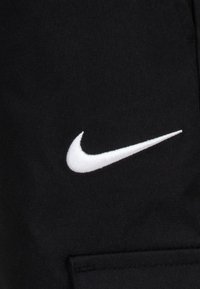 Juodas audinys su tekstūruotu paviršiumi, kuriame matomas didelis baltas siuvinėtas Nike swoosh logotipas kairėje. Minimalistinis dizainas, be papildomų detalių.