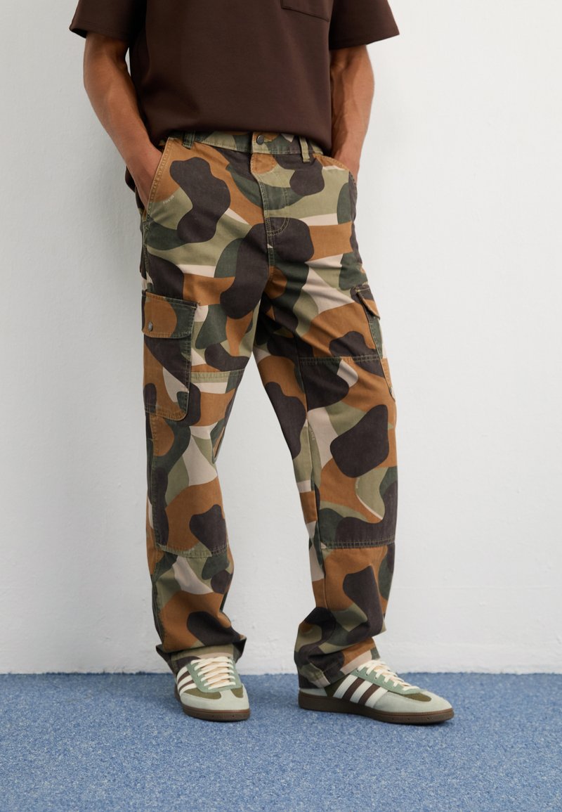 Kamoflage cargo-housut ruskeana, vihreänä ja beigeenä. Sisältää suuret sivutaskut ja rennon leikkauksen. Yhdistetty valkoisiin ja vihreisiin raidallisiin tenniskenkiin.