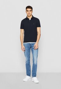Marineblaues Poloshirt mit kurzem Kragen und kleinem Logo, kombiniert mit hellblauen Jeans und weißen Sneakern. Einfaches, lässiges Design.