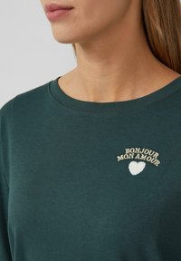 Donkergroene sweatshirt met een ronde hals, voorzien van crèmekleurige geborduurde tekst en een klein hartontwerp op de borst. Zachte textuur.