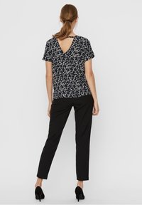 Donna con coda di cavallo marrone, che indossa una blusa nera a maniche corte con motivo e taglio sulla schiena, pantaloni neri aderenti e tacchi neri, in piedi di spalle.