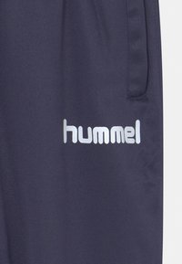 Marina blå atletiska byxor tillverkade av slätt tyg. Har en vertikal ficka och en silverfärgad logotyp på nedre vänstra sidan.