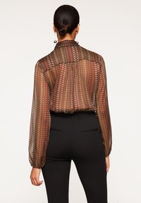 Blusa trasparente in bordeaux e verde con motivo geometrico, con maniche lunghe e colletto semplice, abbinata a pantaloni neri aderenti.