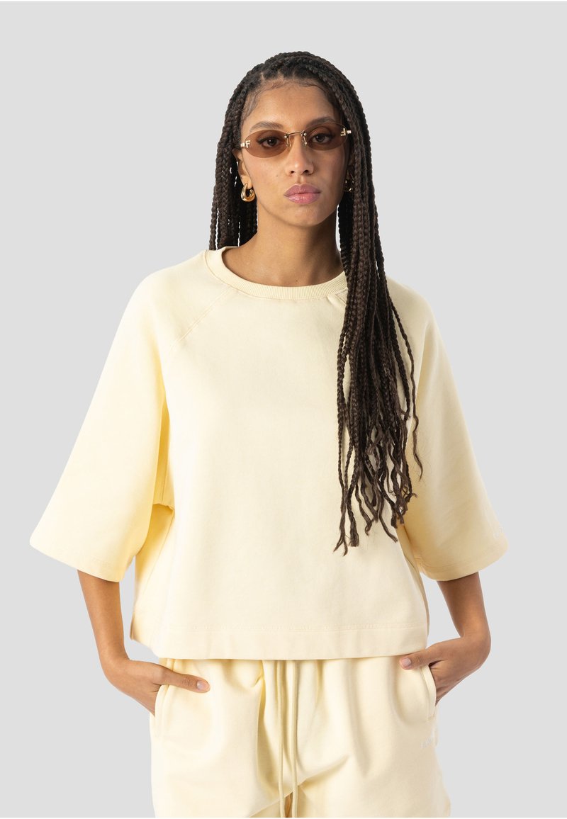 Femme aux longues tresses portant des lunettes de soleil ambrées, un sweat-shirt oversize jaune pâle et un pantalon assorti, les mains dans les poches, fond neutre.