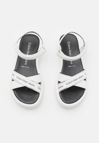 Vita sandaler med en slät ovandel och korsade remmar, svart vadderad innersula och en tryckt logoband, designade i Italien.