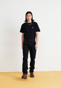 Svart bomull T-skjorte med et lite logo, kombinert med svarte jeans. Figuren har på seg mørkebrune sko og et sølvfarget kjede.
