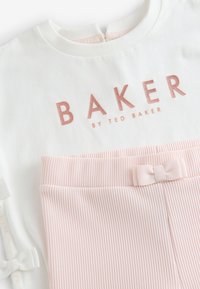 Valkoinen puuvillapaita, jossa on "BAKER" vaalean kultaisella kirjailulla, yhdistettynä vaaleanpunaiseen ribattyyn housuun, jossa on nauha vyötäröllä.