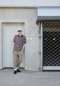 Patterned short-sleeve shirt in black, featuring floral motifs, paired with beige corduroy pants and black slip-on shoes; light blue cap.