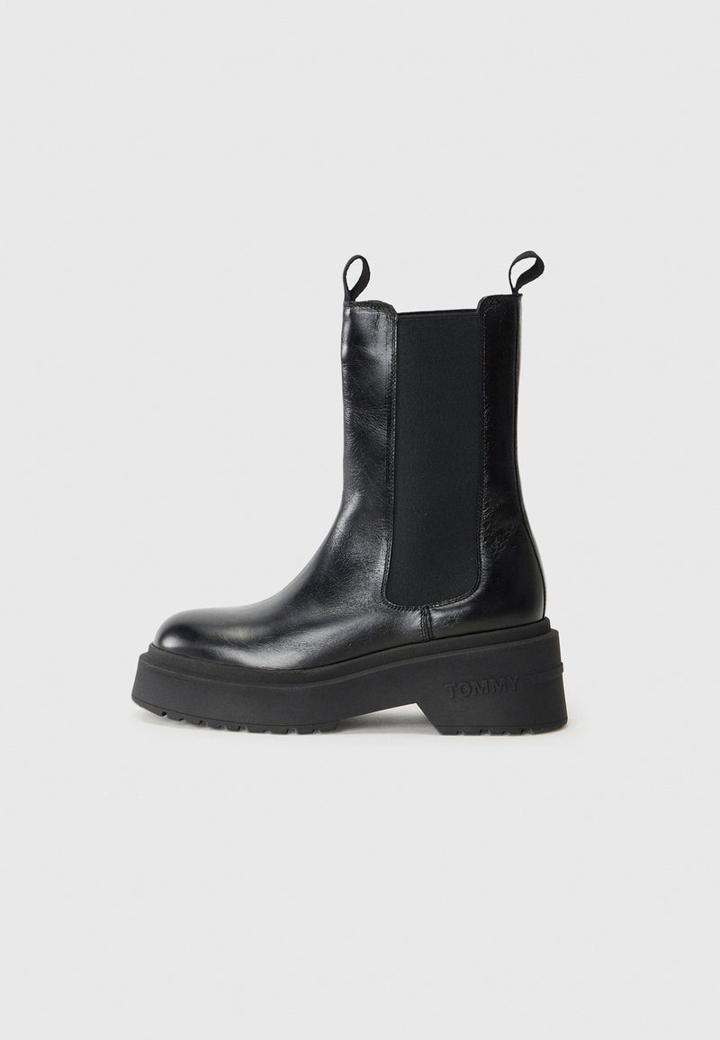Bottine noire en cuir avec semelle épaisse, panneaux latéraux élastiques, languettes de tirage, et "TOMMY" embossé sur la partie talon de la semelle.