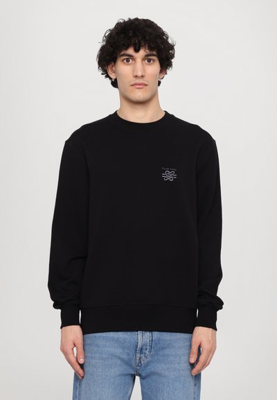 Pull noir à manches longues, col rond et poignets côtelés. Présente un petit logo brodé gris sur la poitrine gauche. Porté sur un jean bleu clair.