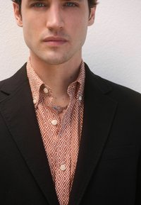 Sacou negru peste o cămașă portocalie cu un model geometric, având butoane albe și un lanț argintiu ca accessorii.