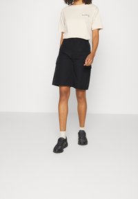 T-shirt beige avec un petit motif, shorts cargo noirs avec poches latérales, et baskets noires avec une surface texturée, portés avec des chaussettes blanches à la cheville.