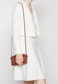 Bílý krčený blazer přes bílé šaty, s malou hnědou koženou kabelkou drženou v levé ruce. Hladká tkanina, minimalistický design.