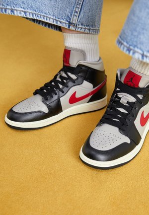 Personne portant des baskets Air Jordan noires, grises et rouges avec des chaussettes blanches et un jean bleu, debout sur un sol jaune moutarde.