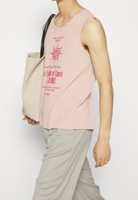 Person wearing a sleeveless pink graphic top and light gray pants, carrying a beige tote bag with black handles over one shoulder.