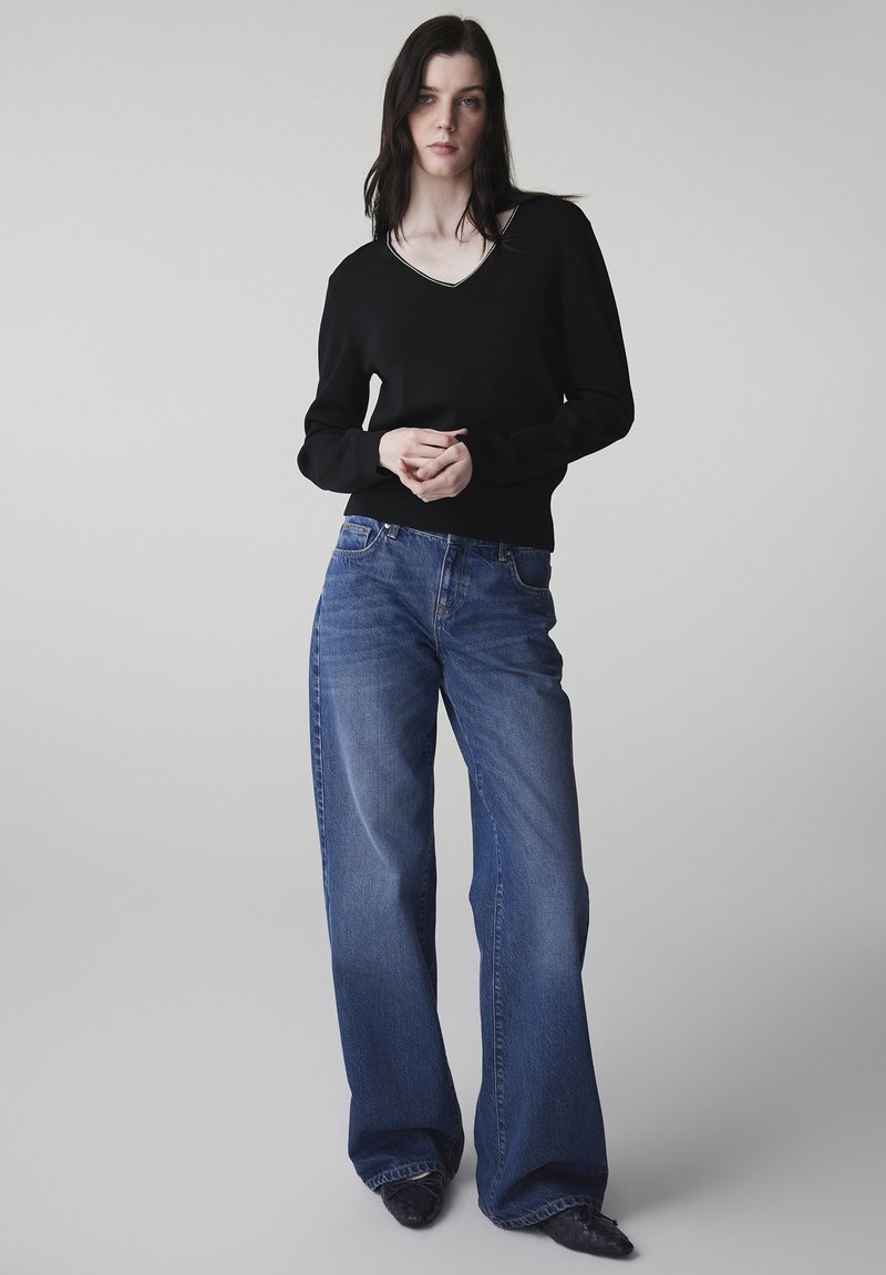 Mujer de pie con una camiseta negra de manga larga y escote en V, jeans azules de pierna ancha y zapatos negros tejidos con punta, sobre un fondo liso.