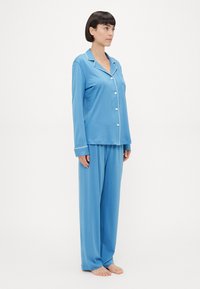 Ανοιχτό μπλε πυτζάμες από μαλακό ύφασμα, με πουκάμισο με κουμπιά και λευκό περίγραμμα, καθώς και παντελόνια σε χαλαρή γραμμή. Απλός, casual σχεδιασμός.