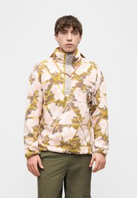 Νέος άνδρας που φοράει ροζ, κίτρινο και καφέ φλις πουλόβερ με σχέδιο φύλλων και ελιά πράσινο παντελόνι cargo, στέκεται μπροστά σε απλό φόντο.