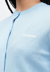 Jasnoniebieski kardigan wykonany z miękkiej dzianiny, z okrągłym dekoltem, niewielkim zapięciem na guziki i subtelnym wytłoczonym logo na piersi.