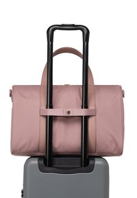 Sac en tissu rose fixé à la poignée d'une valise noire, avec une texture lisse, un dessus arrondi et un accent de fermoir au centre.