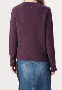 Femme portant un pull à manches longues violet pelucheux et une jupe en denim bleu, vue de dos, debout devant un fond gris clair.