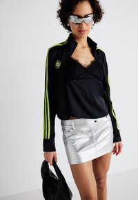 Jachetă sport neagră cu dungi neon verzi, suprapusă peste un top negru cu dantelă, asociată cu o fustă mini metalică argintie și o geantă neagră.