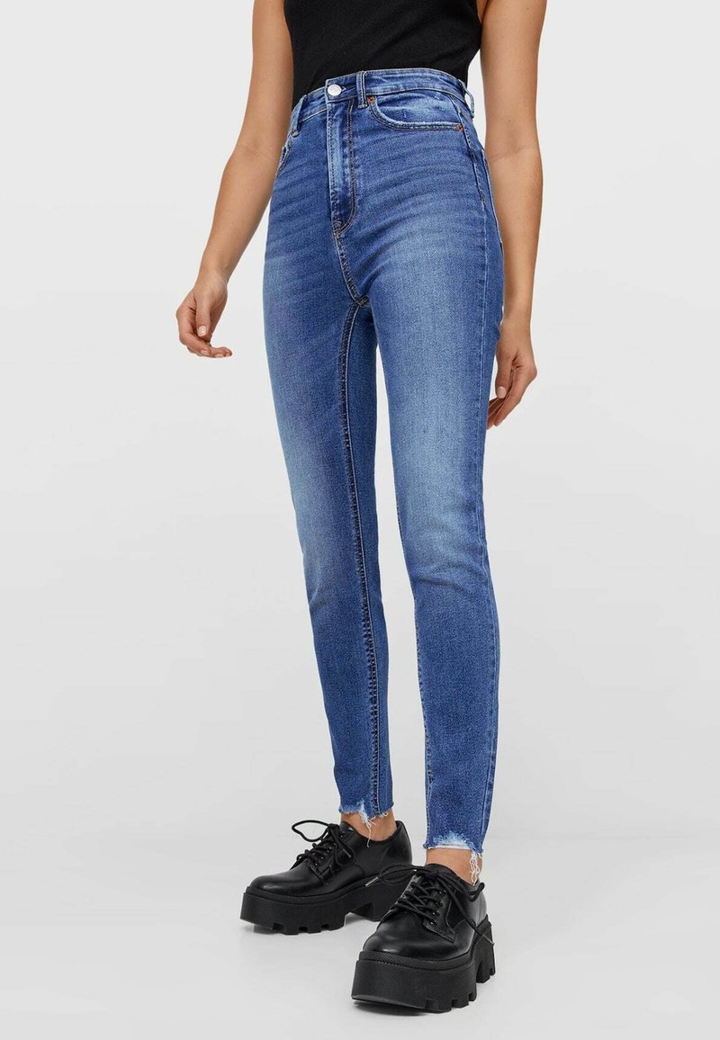 Jean bleu taille haute en denim avec une coupe slim, ourlet brut et deux poches avant, assorti à des chaussures à plateforme noires.
