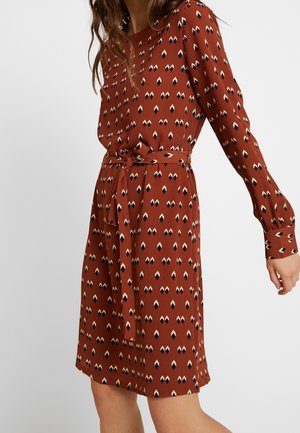 Mujer con un vestido color óxido con patrones de diamantes en blanco y negro y un cinturón atado a la cintura, posando con un brazo ligeramente extendido.