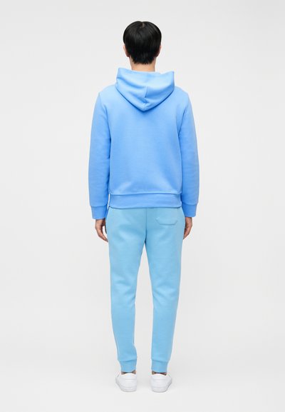 Άνδρας που φοράει ανοιχτό μπλε φούτερ με κουκούλα και αντίστοιχο παντελόνι φόρμας, στέκεται γυρισμένος προς τα πίσω σε απλό λευκό φόντο με λευκά αθλητικά παπούτσια.