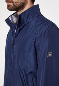 Marineblauw waterdicht jack met een hoge kraag, voorzien van een ritssluiting aan de voorkant, zijzakken en een logopatch op de mouw. Gestreepte voering aan de binnenkant.