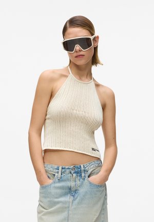 Femme portant des lunettes de soleil à monture blanche surdimensionnée, un haut dos-nu en maille crème, et une jupe en denim bleu clair à taille haute, les mains dans les poches.