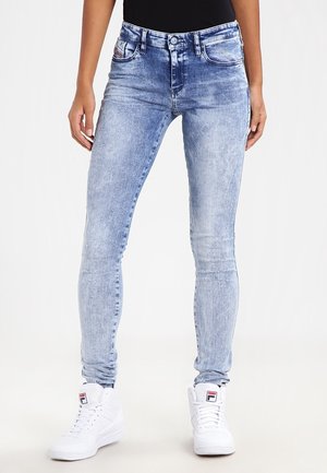 Osoba nosząca jasnoniebieskie obcisłe jeansy acid-wash i białe wysokie sneakersy z logo na jednolitym tle.