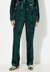 Smaragdgroene fluwelen broek met een gladde textuur, wijde pijpen en elastische tailleband, gecombineerd met opvallende groene hakken.