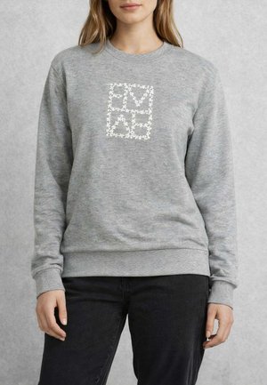 Donna che indossa una felpa grigia con motivo floreale bianco che forma figure quadrate e lettere sul petto, abbinata a pantaloni neri.