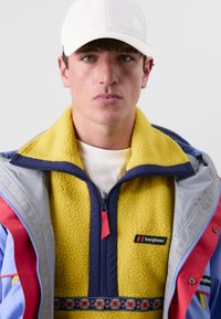 Kleurjack met een gele fleece binnenkant, marineblauwe rits en een bloemenpatroon op de borst. Draagt ook een witte pet.