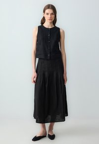 Top senza maniche in lino nero con bottoni frontali, abbinato a una gonna midi plissettata nera. La modella indossa scarpe slip-on nere.
