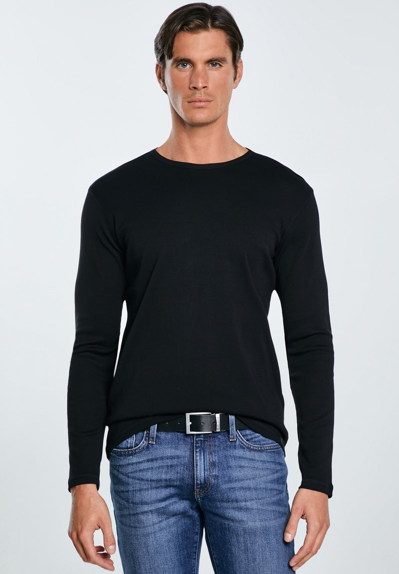 Jaunas vyras su trumpais tamsiais plaukais, vilkintis prigludusią juodą ilgomis rankovėmis marškinių, mėlynų džinsų ir juodą diržą, stovintis priešais vientisą foną.