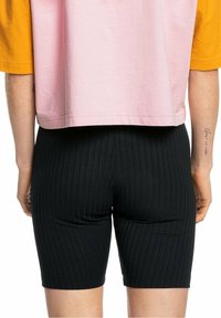 Černé žebrované šortky mají těsný střih, které jsou kombinovány s krátkým růžovým a žlutým topem. Design zdůrazňuje vertikální texturu a pružnost.