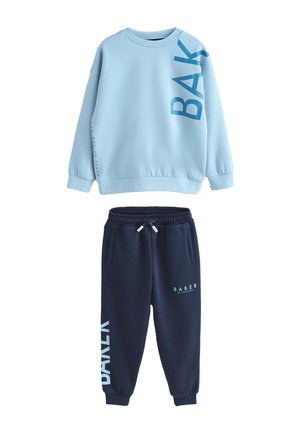 Junge steht in hellblauem übergroßem Sweatshirt mit dem Schriftzug "BAY", navyblaue Jogginghose mit dem Schriftzug "BAKER" und weißen Turnschuhen auf grauem Boden und Wand.