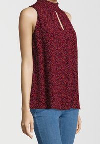 Sleeveless red blouse with black leopard print, keyhole neckline, worn with blue jeans, shown from the side on a standing person.