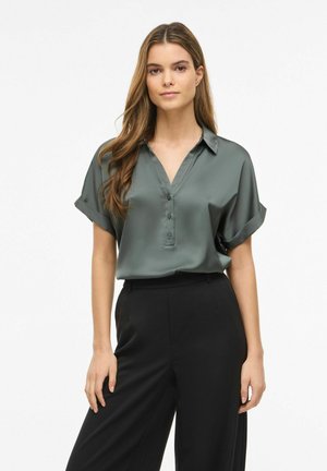 Jeune femme aux longs cheveux portant un chemisier vert en satin à manches courtes et un pantalon noir taille haute, debout sur un fond blanc uni.