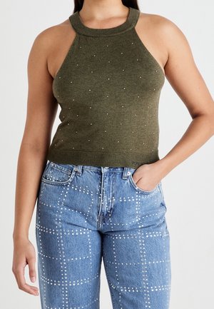 Femme portant un haut en tricot sans manches vert olive avec de petits détails en perles et un jean bleu orné d'un motif en grille avec des clous argentés.