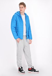 Giacca a vento blu con cappuccio, texture trapuntata, indossata sopra una maglietta bianca e pantaloni della tuta grigi, completata da sneakers nere e rosse.