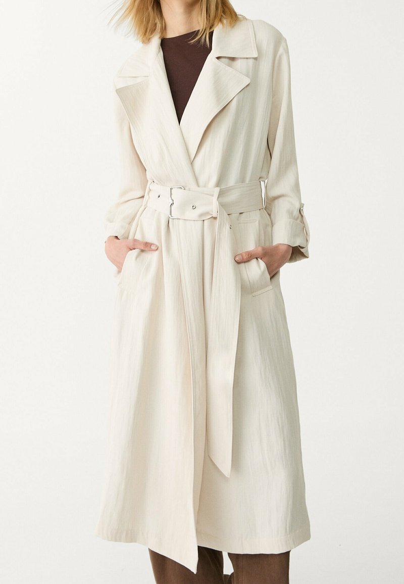Femme portant un long trench-coat crème ceinturé avec de larges revers et les mains dans les poches, par-dessus un top sombre et un pantalon marron.