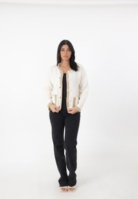 Cardigan crema con rifiniture e bottoni beige, dotato di due tasche. Abbinato a pantaloni neri e sandali bianchi con punte scoperte, su uno sfondo neutro.