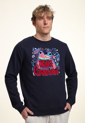 Jonge man die een marineblauwe trui draagt met een kleurrijk cartoonfiguur en de tekst "Weird and Complicated" op de voorkant, staand met handen in de zakken.