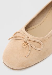 Μπεζ σουέντ μπαλαρίνα με στρογγυλή μύτη, διακοσμητική κορδέλα και χρυσό στοιχείο. Μαλακή υφή και ελάχιστες ραφές.