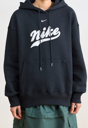 Person trägt einen schwarzen Nike-Hoodie mit weißem Logo und grüne Cargo-Hose, steht vor einem schlichten hellen Hintergrund.