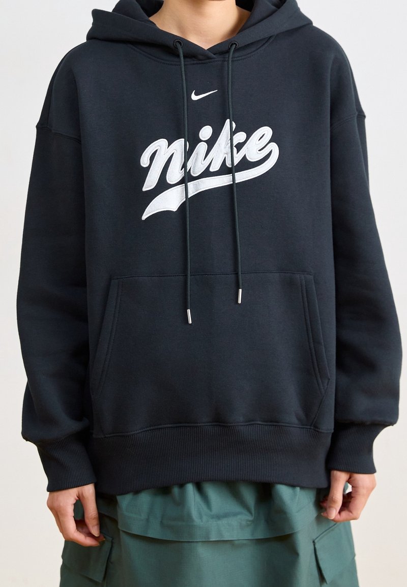 Person klädd i en svart Nike-ziphoodie med vitt logotyp och gröna cargobyxor, stående mot en ljus, enfärgad bakgrund.